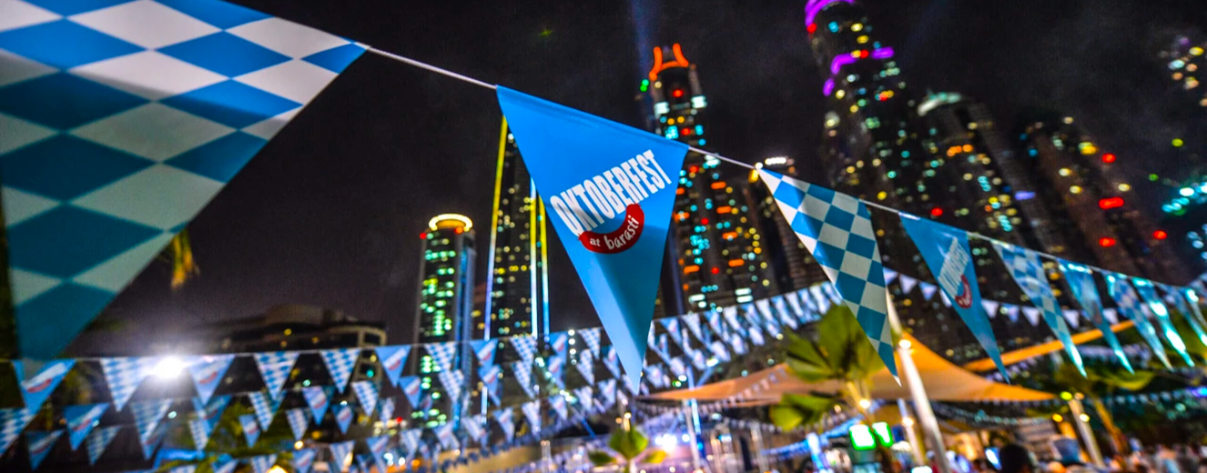 Blue Oktoberfest flag with white lettering, illuminated by vibrant city lights at night.