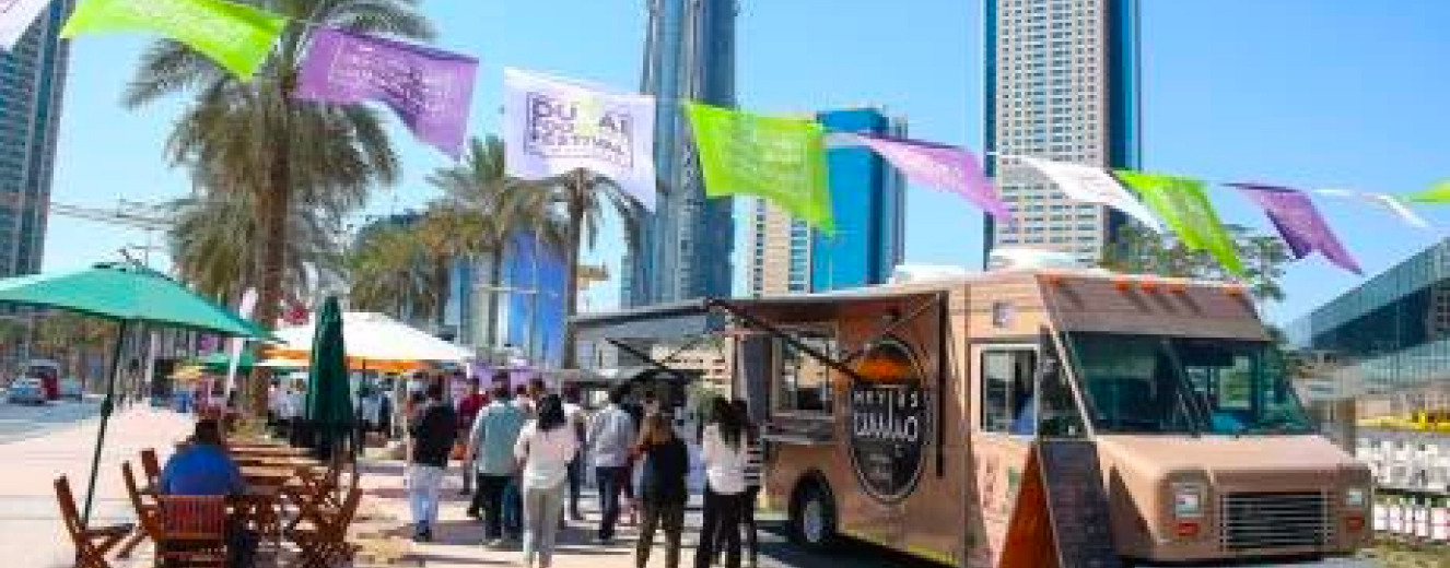Crowd gathered around a food truck, with colourful banners and tall buildings in the background.