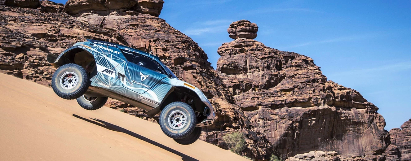 A blue and white off-road vehicle climbs a sandy dune against a backdrop of rocky formations.