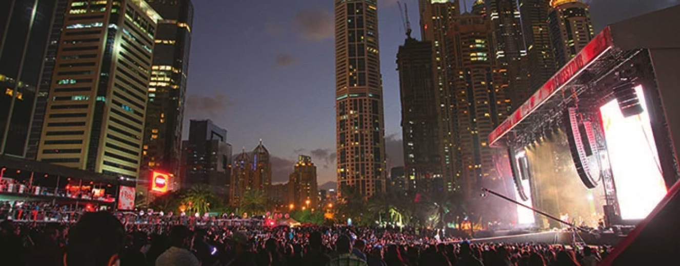 Crowd illuminated by colourful lights at dusk, with tall buildings in the background.