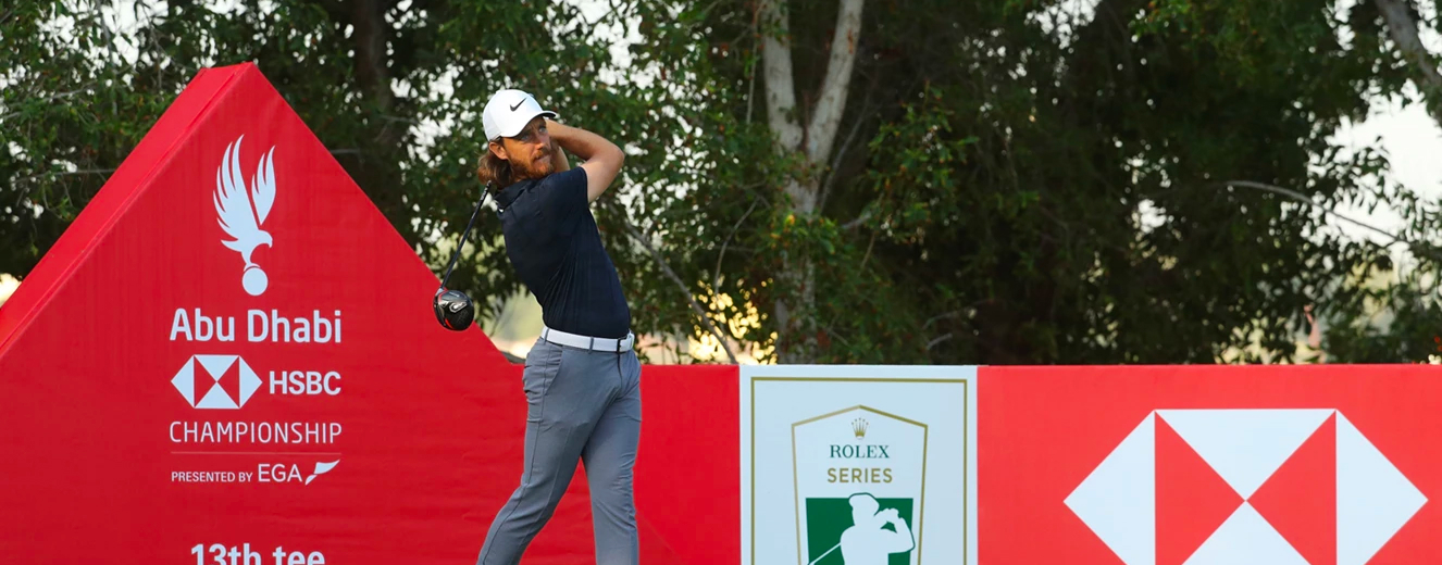 Golfer in a navy shirt and grey trousers swings a club near a green and red sign.