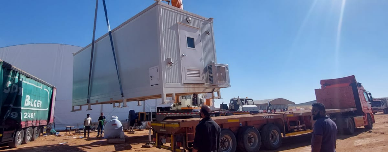 A white temporary kitchen unit is being lifted onto a truck against a clear blue sky.