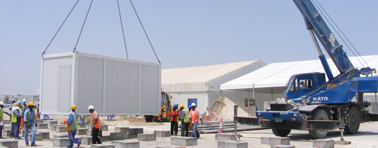Construction workers in hard hats oversee a container being lifted at a temporary kitchen site.