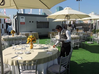 A waiter in a black vest arranges a table with a yellow floral centerpiece under a sun umbrella.