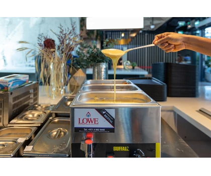 A hand pours a creamy sauce from a ladle into a stainless steel serving container.
