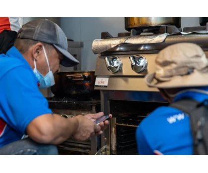 Two individuals in blue shirts examine an oven, with stainless steel appliances in the background.