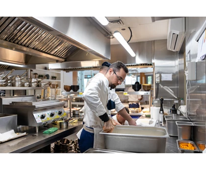Two chefs in a stainless steel kitchen, one stirring a large pot, surrounded by cooking equipment.