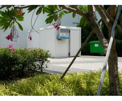 A white utility shed surrounded by green foliage and pink flowers, with a green bin nearby.