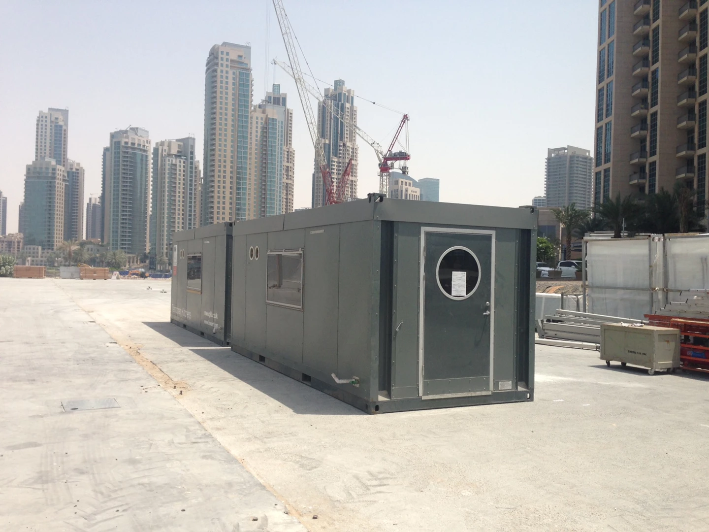 Grey construction container on a sandy site, with tall buildings and cranes in the background.