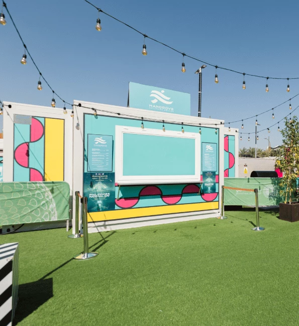 Brightly coloured food kiosk with a turquoise backdrop, surrounded by green grass and decorative string lights.