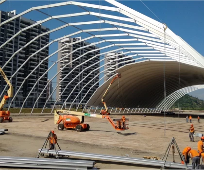 Construction site featuring a white steel frame structure under a clear blue sky and nearby buildings.
