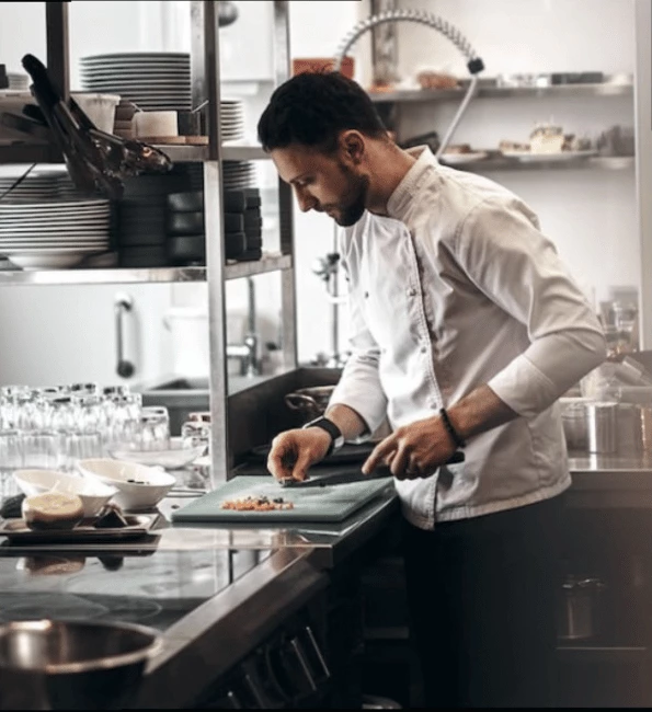 A chef in a white uniform prepares food on a green cutting board in a modern kitchen.