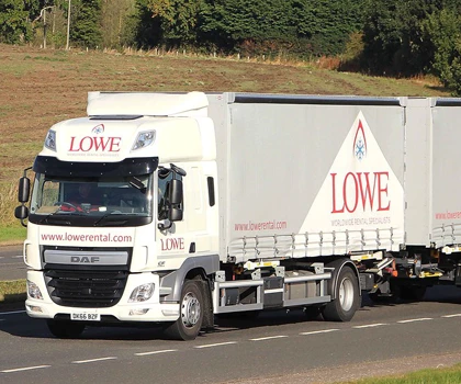 White delivery truck with "LOWE" branding driving on a rural road, surrounded by green fields.
