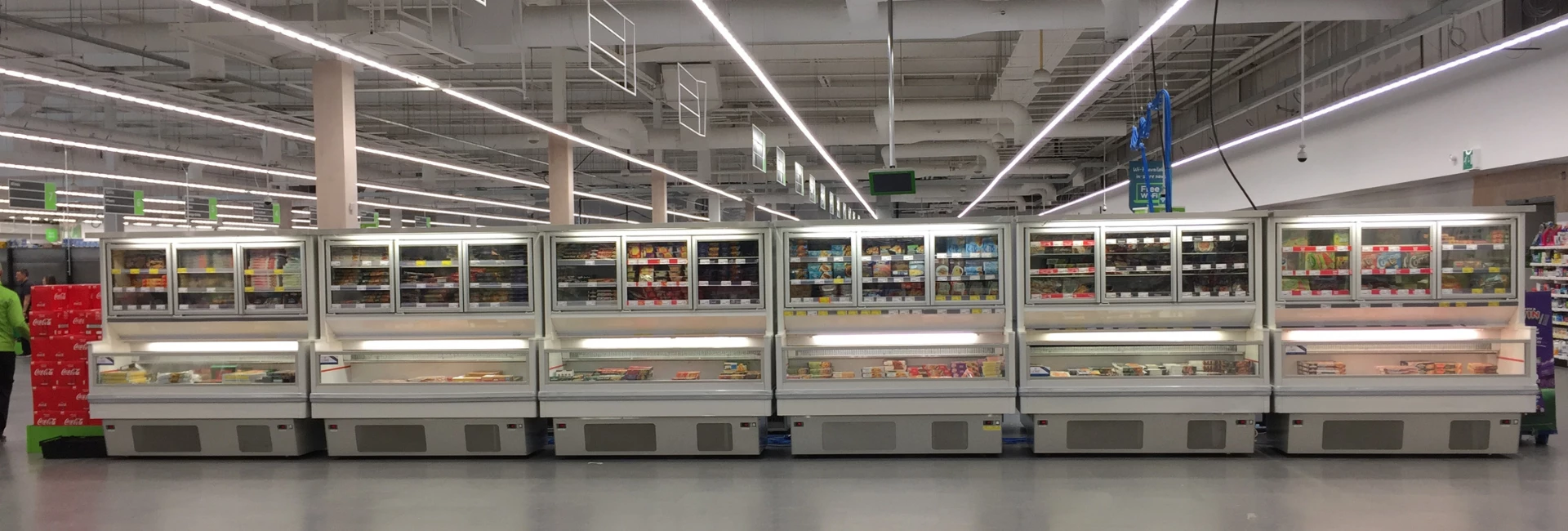 Brightly lit supermarket aisle featuring multiple refrigerated units filled with various packaged products.