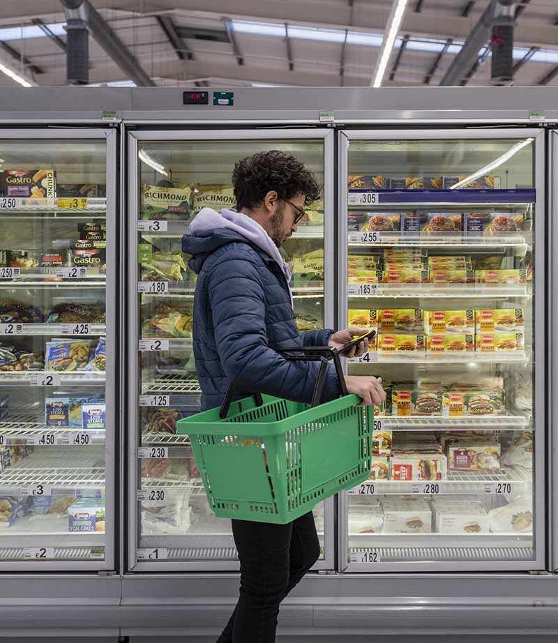 A person in a blue jacket holds a phone while standing near a freezer with colourful food packaging.