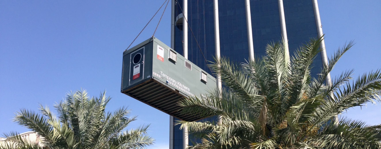 Green shipping container suspended above palm trees, with a modern building in the background.