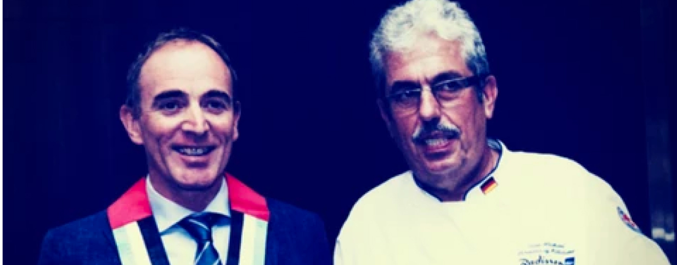 Two men smile, one in a dark suit with a red sash, the other in a white chef's coat.