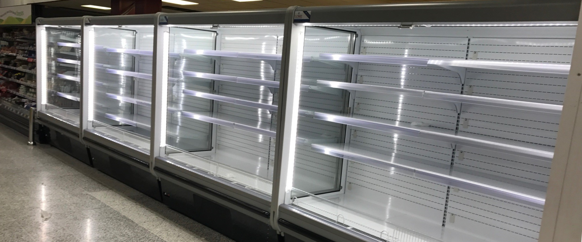 Empty commercial refrigerator with sleek silver shelves and bright white lighting, set against a neutral background.