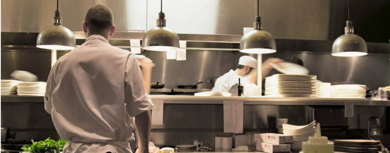 Chefs in white uniforms work in a busy kitchen, with metallic surfaces and warm lighting.