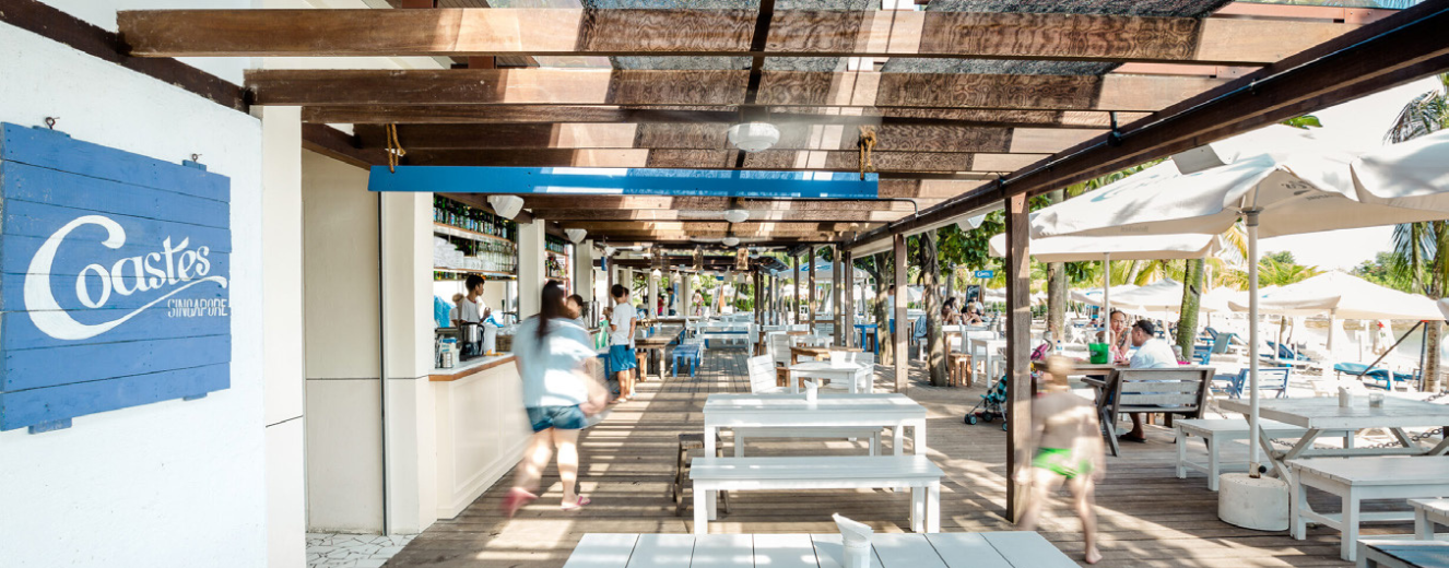 Bright, open dining area with white wooden tables and shaded seating under a wooden pergola.
