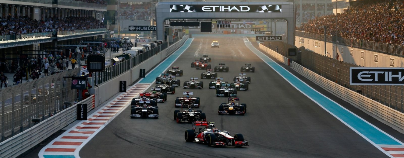 Formula 1 cars racing on a circuit, with a blue track and an illuminated grandstand in the background.