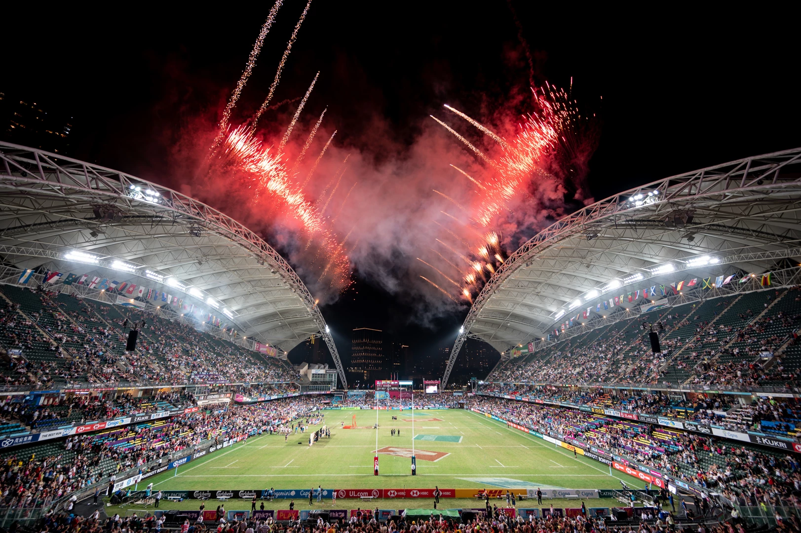 Fireworks illuminate a stadium at night, with a vibrant crowd watching a rugby match below.