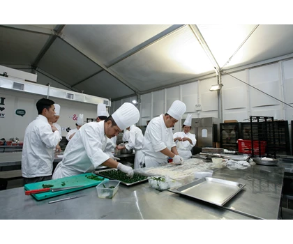 Chefs in white uniforms and hats prepare dishes in a well-lit tent during the festival.