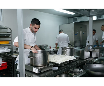 Chefs in white uniforms prepare food in a modern kitchen, with stainless steel pots and equipment.