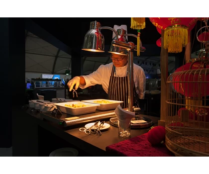 A chef in a striped apron prepares food under warm lighting, with decorative red elements in the background.