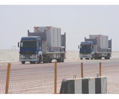 Three blue trucks transport cargo along a sandy road, with a barrier visible in the foreground.