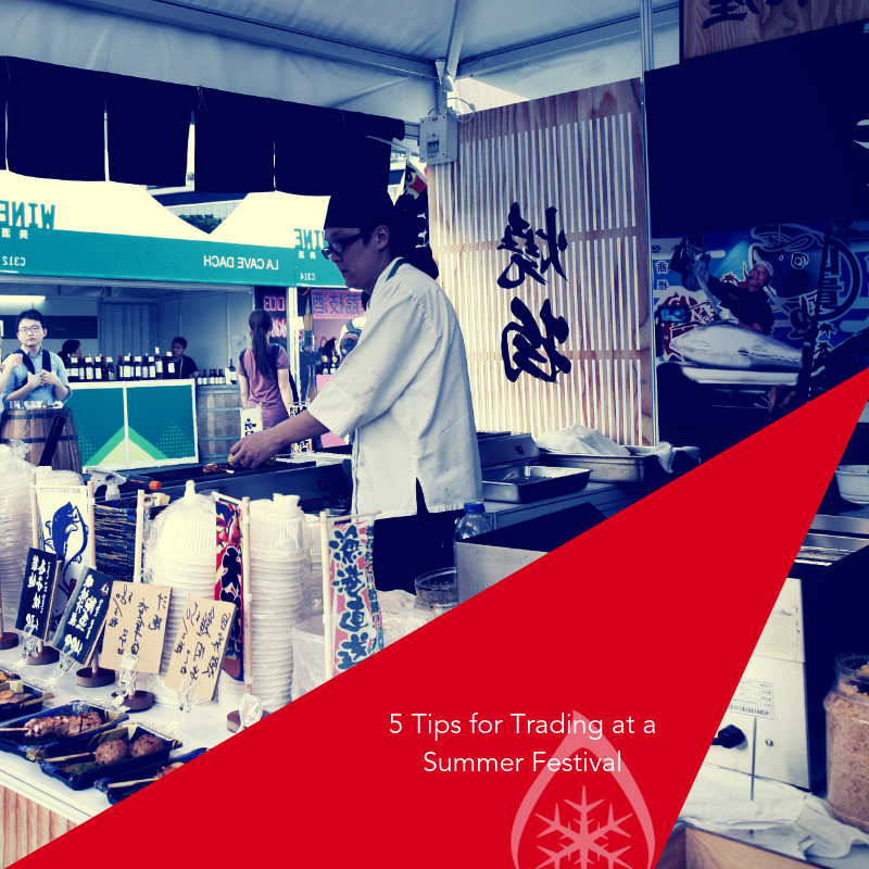 A chef prepares food at a summer festival stall, surrounded by colourful signage and ingredients.