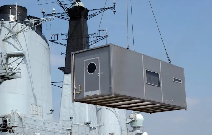 A grey shipping container is being lifted by a crane against a clear blue sky.