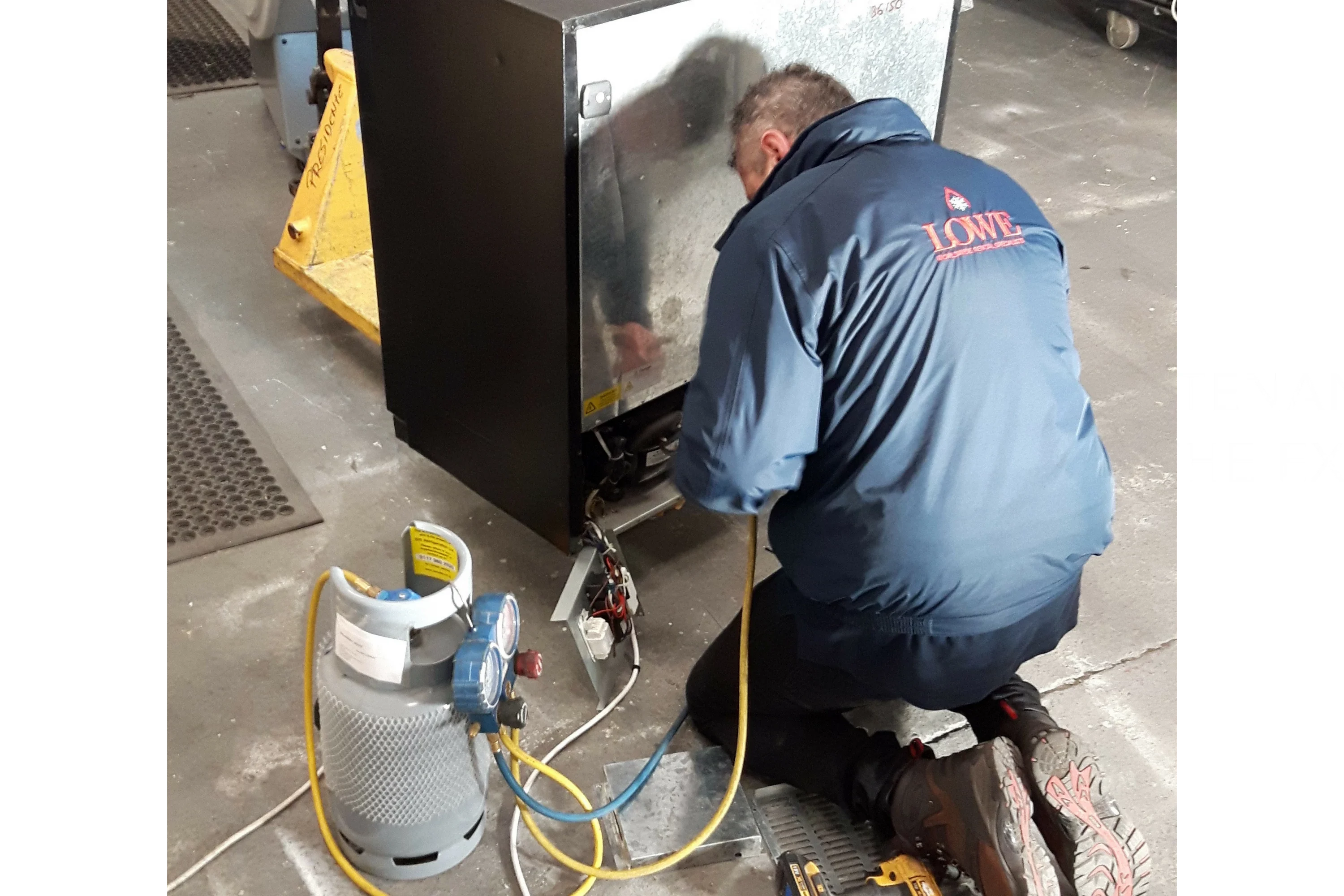 A technician in a blue jacket kneels beside a black appliance, connecting a gas line.