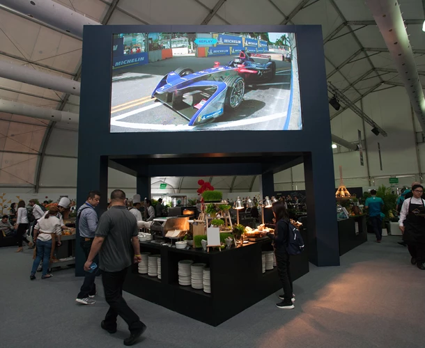 A large screen displays a blue and red racing car, surrounded by event attendees in a tent.
