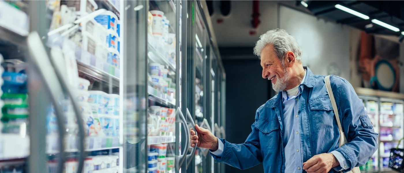 An older man with grey hair smiles while reaching for a product in a glass refrigerator.