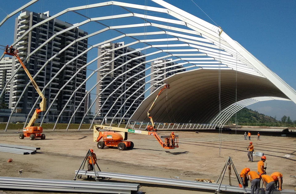 Steel framework of a large structure under construction, with blue sky and nearby buildings in background.