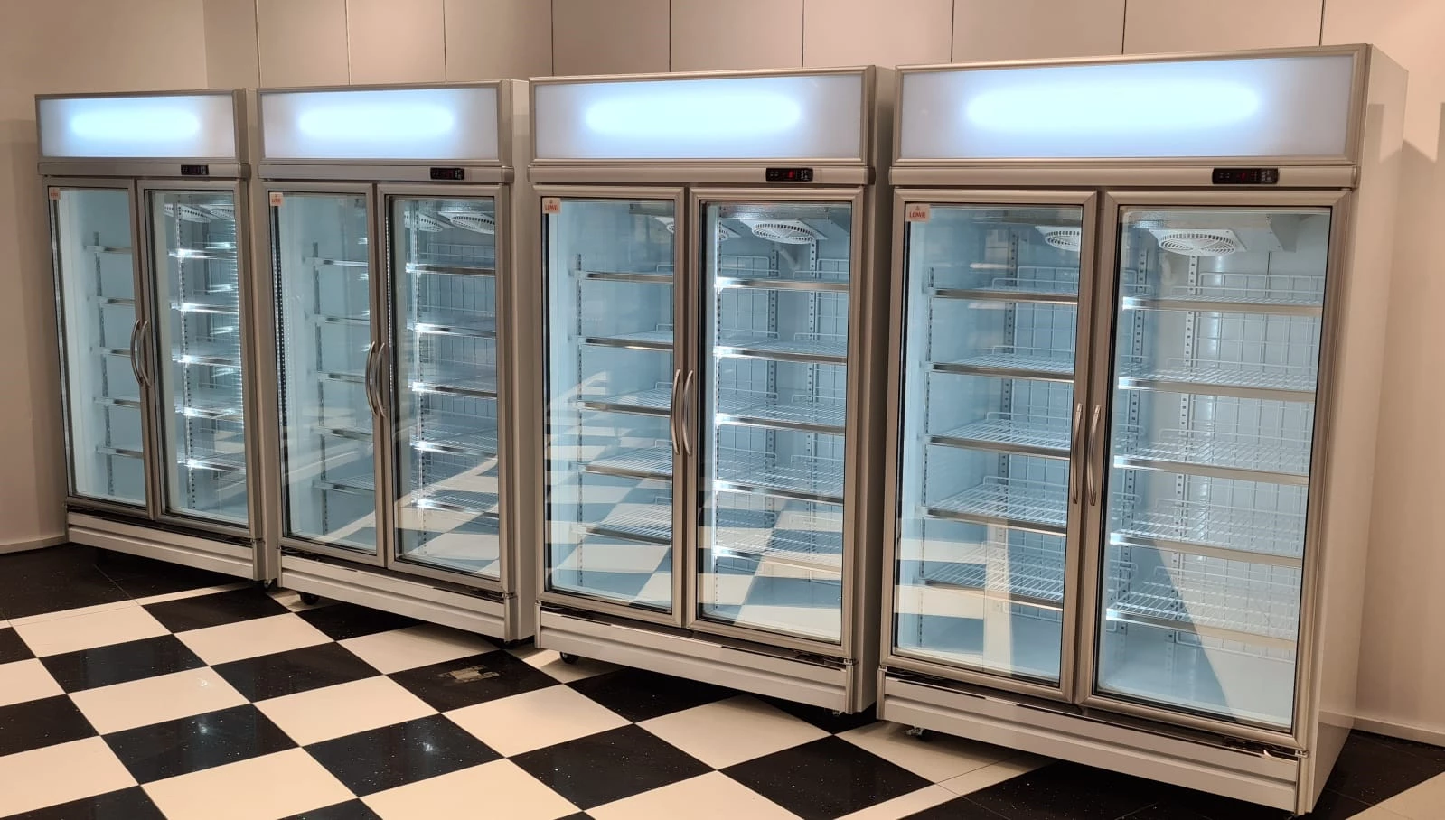 Four glass-fronted refrigerators with empty shelves, set against a black and white tiled floor.