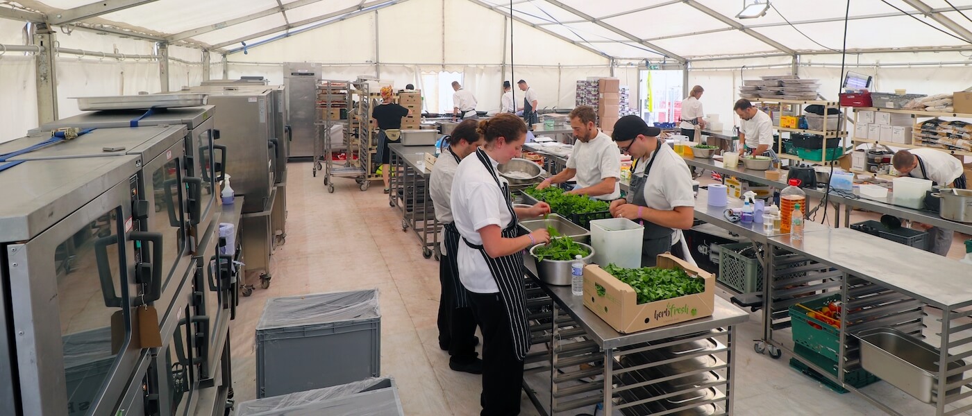 Chefs in white uniforms prepare fresh greens in a large, well-lit kitchen tent.