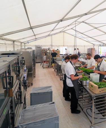 Catering staff in a tented kitchen prepare fresh ingredients on stainless steel tables.