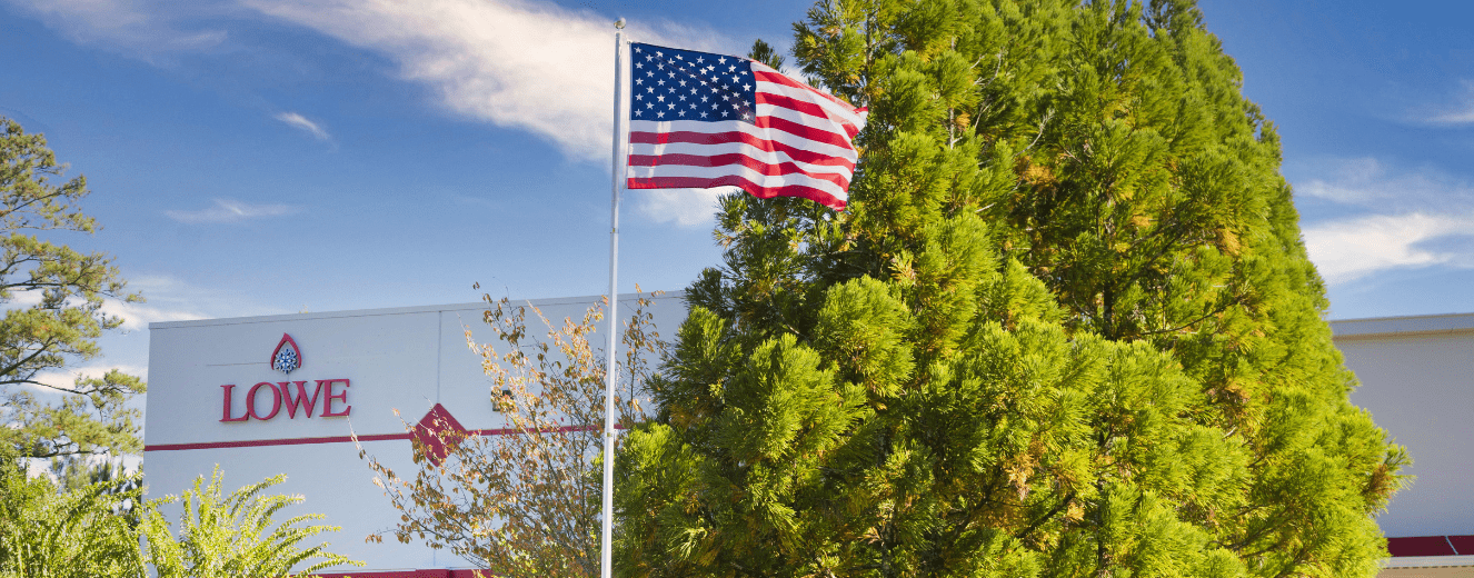American flag waving against a clear blue sky, with green foliage in the background.
