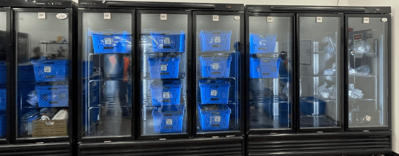 Blue grocery pickup bins stacked inside glass-fronted refrigeration units in a retail environment.