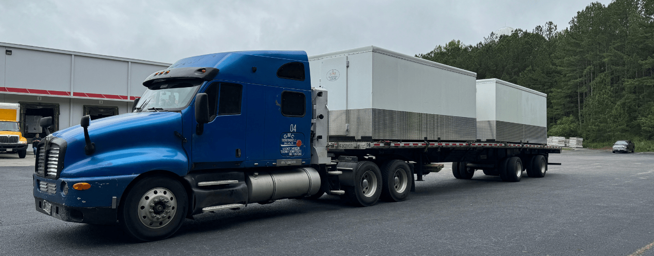 A blue truck transporting a large white walk-in cooler on a grey road under cloudy skies.