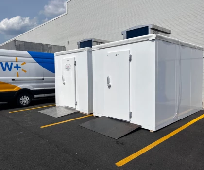 Two white storage units with ramps parked on a tarmacked area beside a delivery van.