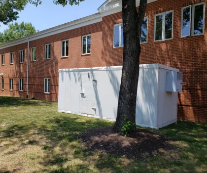 White storage unit beside a tree, with a brick building and green grass in the background.