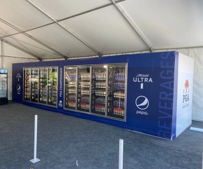 Beverage vending area featuring glass-fronted coolers against a blue backdrop, inside a tent.