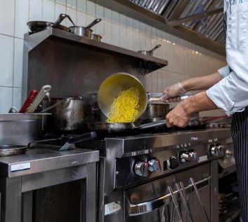 A chef pours yellow pasta into a pot on a stainless steel stove in a commercial kitchen.