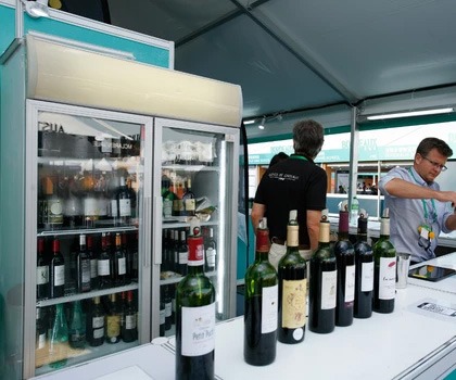 A selection of red wine bottles lined up on a table, with a glass-fronted fridge in the background.