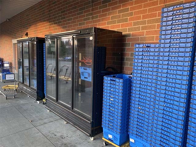 Black glass-fronted refrigerators beside a stack of blue plastic crates against a brick wall.