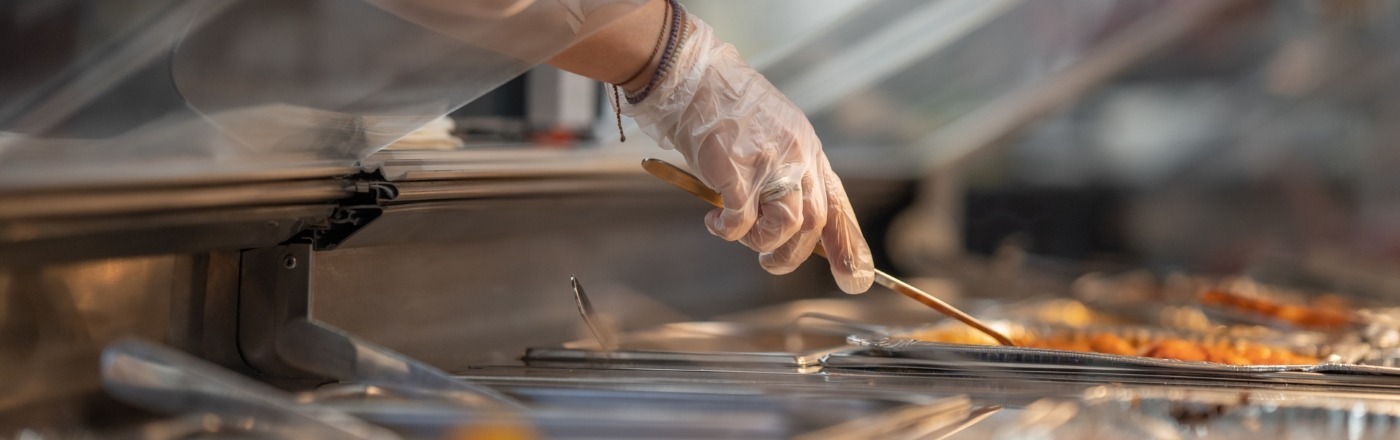 A gloved hand uses a wooden spatula to serve food from a stainless steel container.