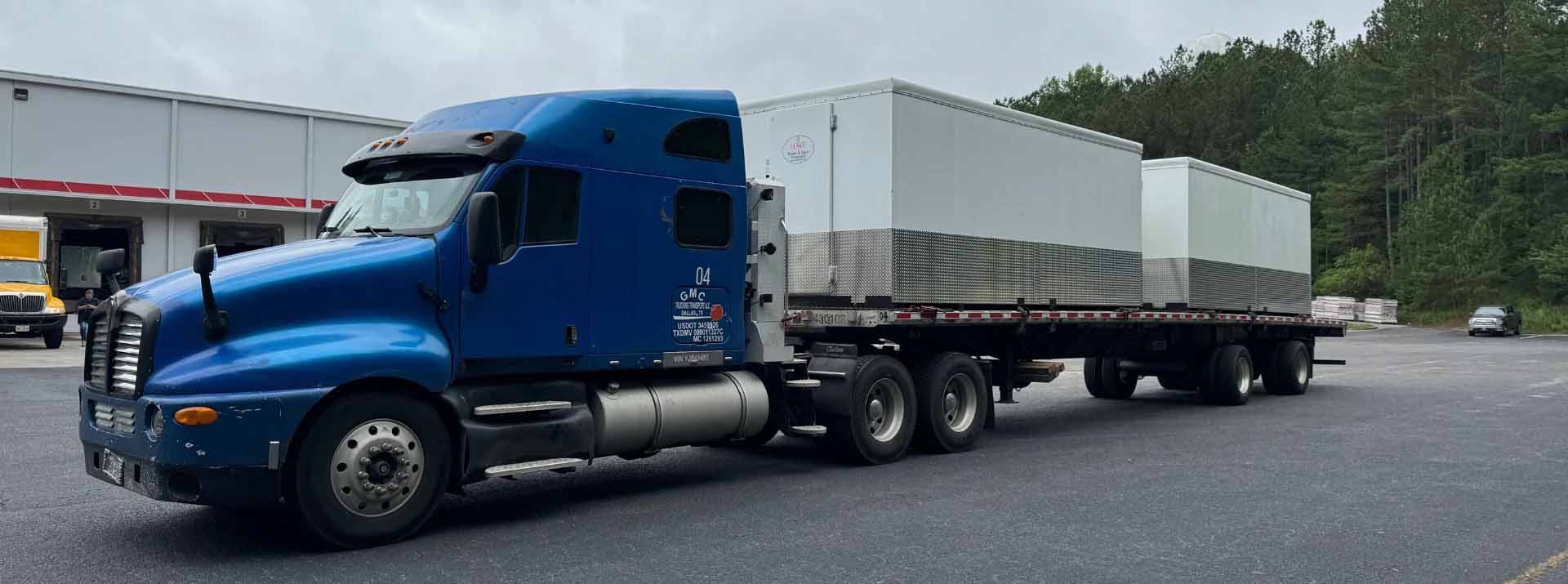 A blue truck transporting a large white container on a flatbed trailer.